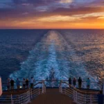 석양으로 붉게 물든 바다 위를 항해하는 크루즈 선박의 후미 갑판. 승객들이 난간에 기대어 샴페인 잔을 들고 수평선을 바라보고 있으며, 배 뒤로는 하얀 물살이 길게 이어져 있다.